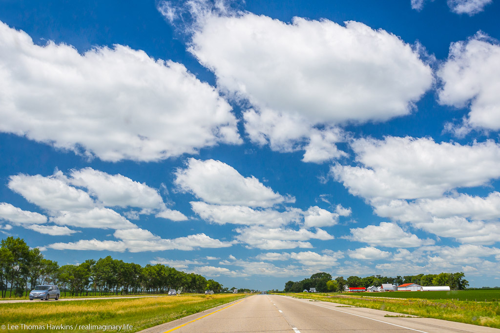 Cumulus clouds stretch across the sky over I-90 and prairie farmlands near Blue Earth, Minnesota.