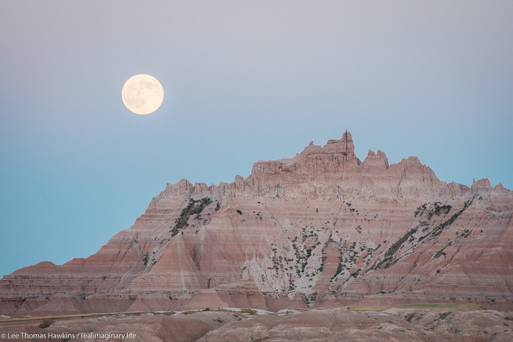 A full moon rises over The Castle at sunset from the White River Valley Overlook in Badlands National Park in South Dakota.