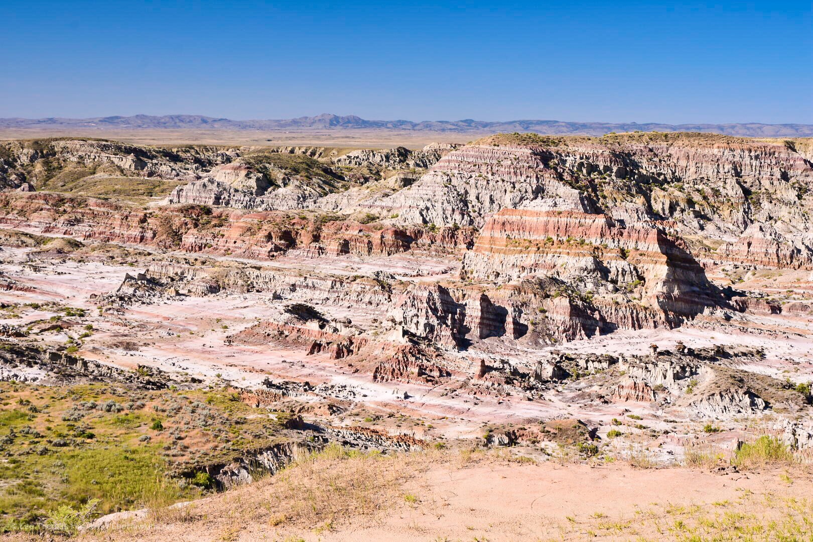 A multilayered jagged rock wall in a canyon with mountains in the distance against a blue sky