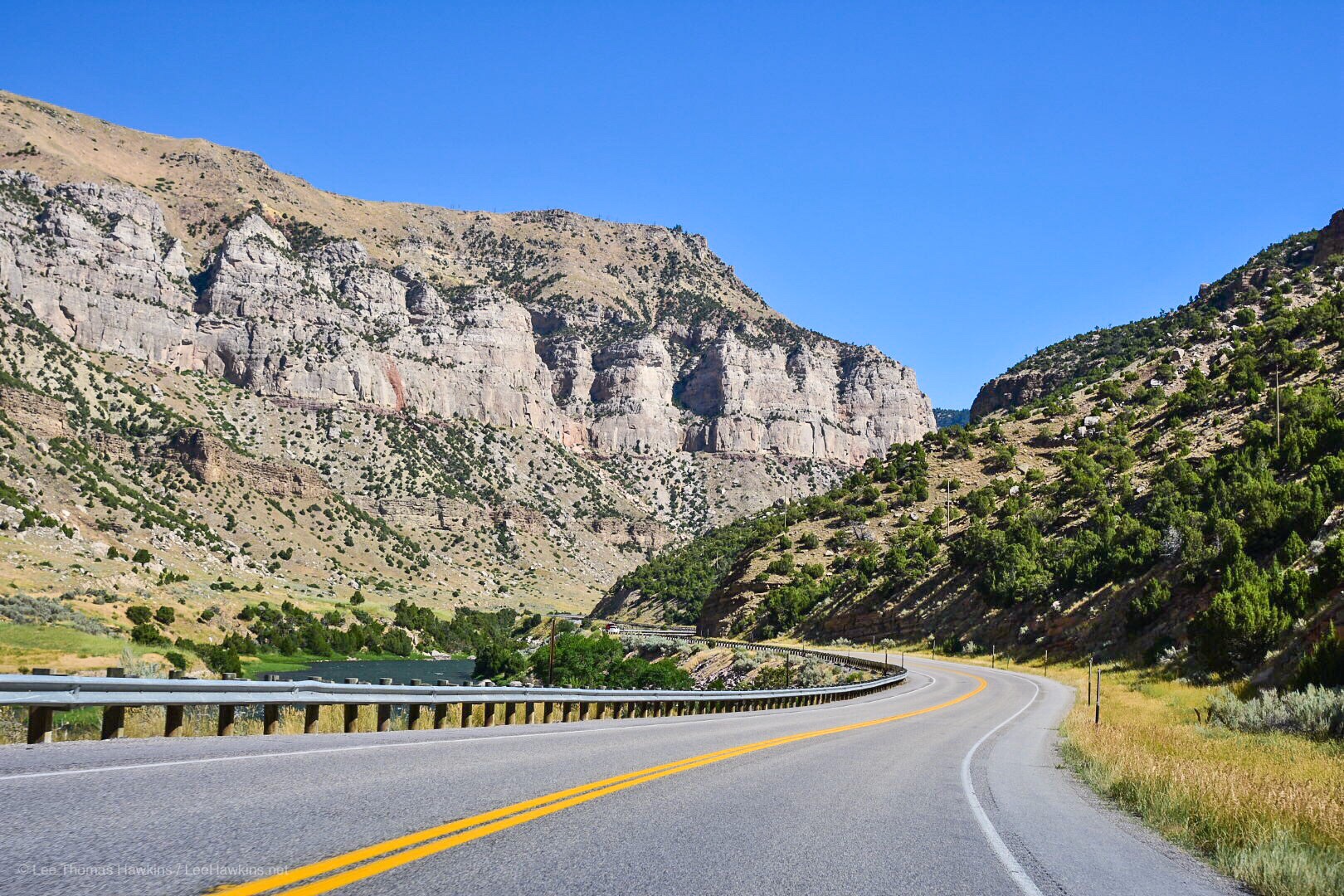 A mountain rises with a sheer rock face high above a canyon, with a shorter rock face rising to the right. A river flows through the center and cars move along a road just above and to the right of it.