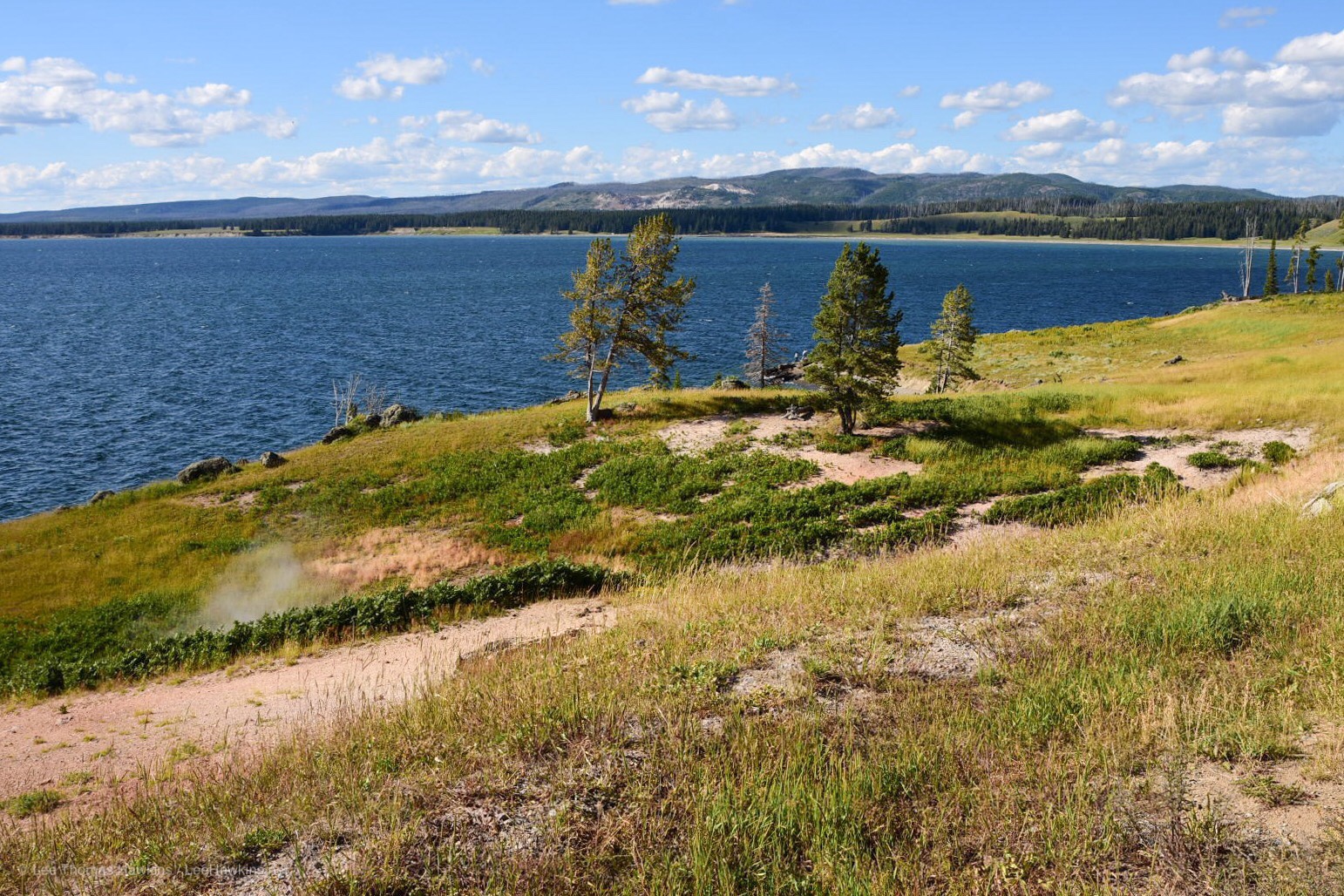 Steam rises from a fumarole along the steep shore of a large blue lake with mountains and a partly cloudy blue sky in the distance.