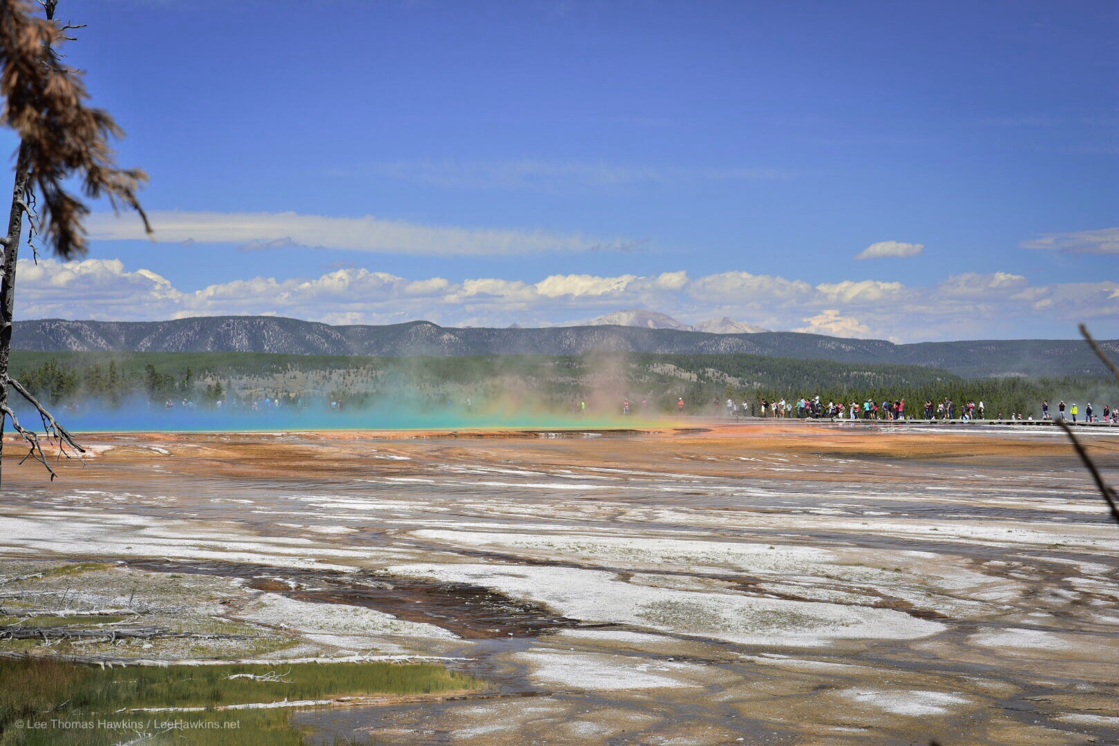 Colored steam rises from a hot spring in a barren landscape surrounded by picturesque mountains.