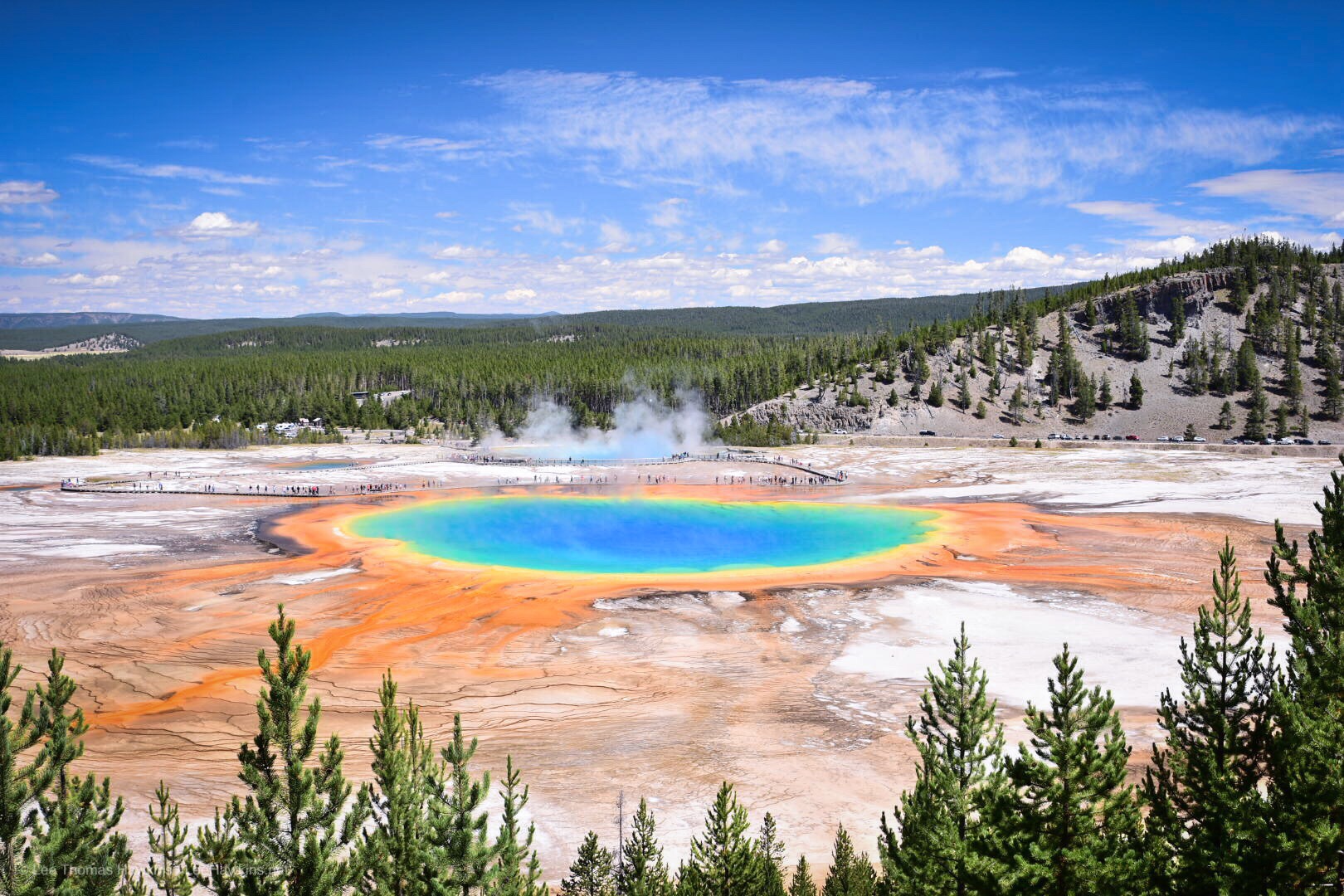 A gigantic hot spring with deep blue and green colors in the center is surrounded by yellow, orange, and red colors on the fringes.