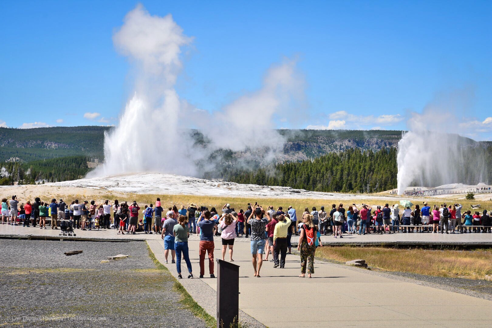 Two large geysers erupt simultaneously, one in the foreground surrounded by a crowd of spectators, and the other in the distance.